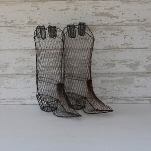 May include: A pair of decorative wire cowboy boots. The boots are made of dark brown wire and have a textured appearance. The boots are standing upright on a white surface, with a distressed white wooden background.