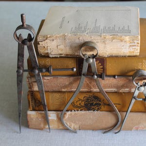 May include: Three antique metal calipers of varying sizes stand before a stack of aged books with yellowed pages. The books have gold-toned covers and visible wear, suggesting age and use. The calipers are silver-toned and show signs of age.