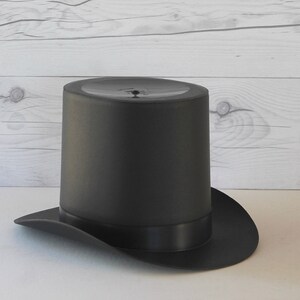 May include: A black top hat with a wide brim. The hat is made of a smooth, dark fabric.