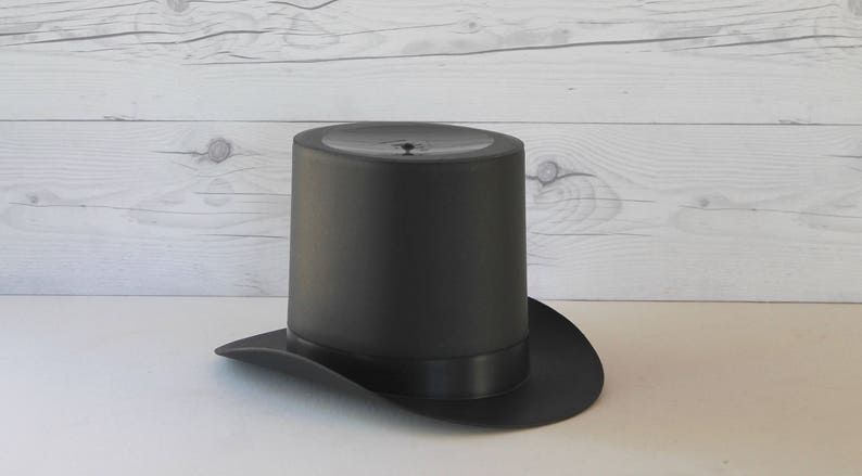 May include: A black top hat with a wide brim. The hat is made of a smooth, dark fabric.