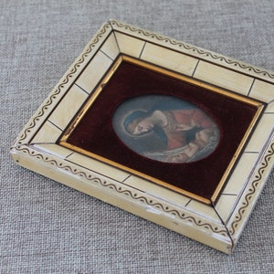 May include: A small, framed artwork featuring a painting of a woman holding a baby. The frame is made of ivory-colored wood with a decorative border. The painting is set against a burgundy velvet background.