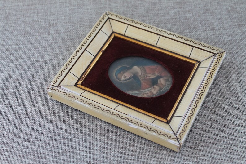 May include: A small, framed artwork featuring a painting of a woman holding a baby. The frame is made of ivory-colored wood with a decorative border. The painting is set against a burgundy velvet background.