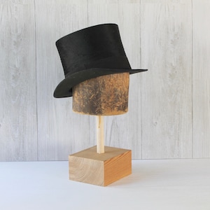 May include: A black top hat on a wooden hat stand. The stand has a square base and a round top.