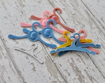 May include: A collection of plastic doll hangers in various colors, including blue, pink, and yellow. The hangers feature a child's face on the top hook. Some of the hangers have the text "Dolly's Pride" printed on them.
