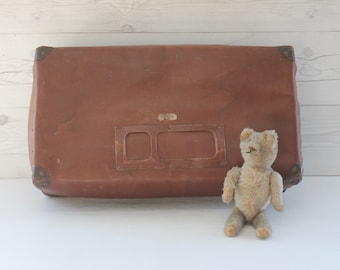 May include: A vintage brown leather suitcase with two rectangular metal name tag holders and a small, worn, beige teddy bear sitting in front of it.
