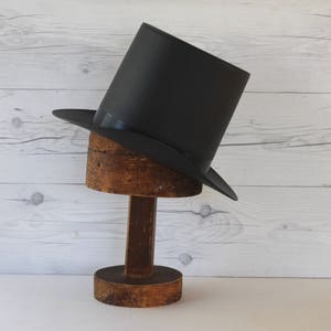 May include: A black top hat on a wooden hat stand. The hat is tilted to the side and the stand is made of rough-hewn wood.
