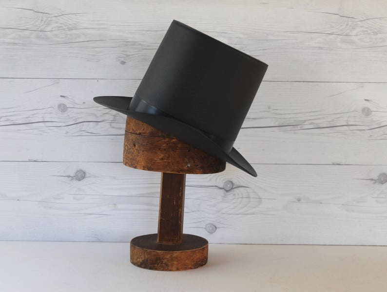 May include: A black top hat on a wooden hat stand. The hat is tilted to the side and the stand is made of rough-hewn wood.