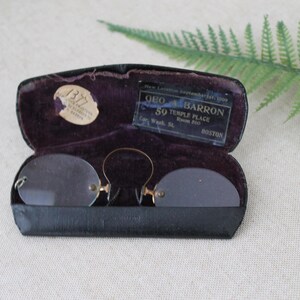 May include: A black leather case with a purple velvet interior holds a pair of antique gold-rimmed glasses. The case has a label that reads "GEO. A. BARRON 59 TEMPLE PLACE BOSTON".