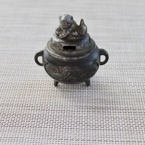 May include: A small, silver-colored incense burner with a lion-shaped lid. The burner has intricate floral designs and three small feet.