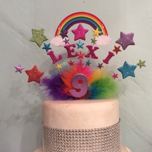 May include: A pink and white cake with a rainbow, stars, and the name "Lexi" on top. The cake is decorated with a silver glitter band and a pink number 9.