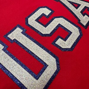 Könnte beinhalten: Roter Pullover mit dem Wort "USA" in großen, weißen, glitzernden Buchstaben, die mit Blau umrandet sind.