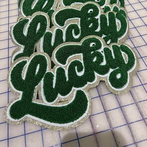 Puede incluir: Una pila de parches "Lucky" verdes y dorados con un contorno blanco. Los parches están hechos de un material peludo y tienen un acabado brillante.