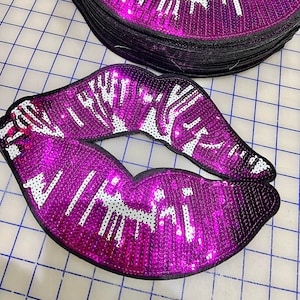 May include: A stack of purple and white sequined lips patches. The patches are made of sequins and have a black backing.