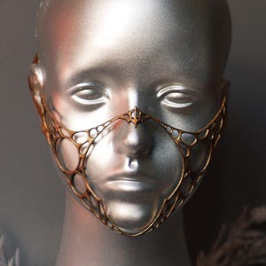 Könnte beinhalten: Eine goldfarbene Filigran-Gesichtsmaske mit kreisförmigem Muster, präsentiert auf einem silbernen Schaufensterkopf. Die Maske bedeckt Nase und obere Wangen. Der Text "Black Unicorn" befindet sich unten.