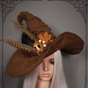 Puede incluir: Un sombrero de bruja marrón con una corona puntiaguda, decorado con plumas, setas y una hoja. El sombrero lo lleva puesto un maniquí con el pelo largo y plateado.