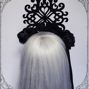 Puede incluir: Diadema negra con un gran dise&ntilde;o de corte negro intrincado y dos acentos de flores negras con centros verdes. La diadema se lleva en una modelo con el pelo largo y blanco.