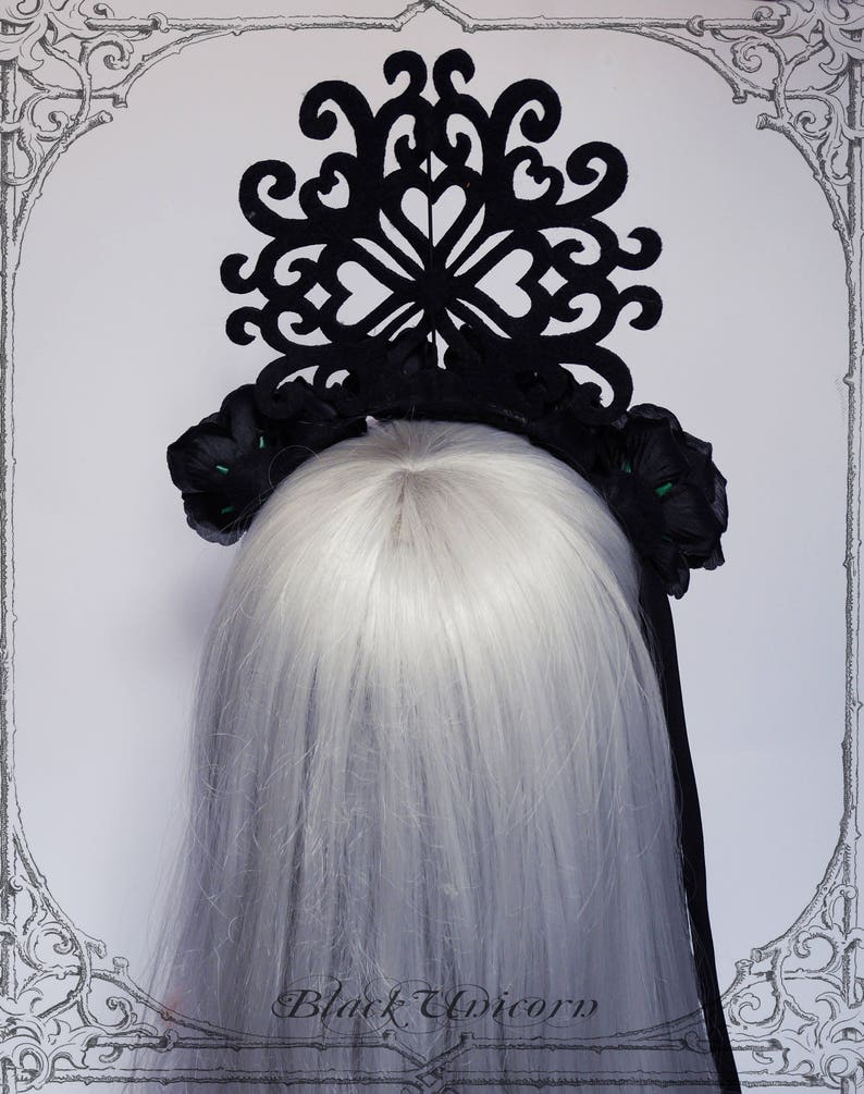 Puede incluir: Diadema negra con un gran dise&ntilde;o de corte negro intrincado y dos acentos de flores negras con centros verdes. La diadema se lleva en una modelo con el pelo largo y blanco.