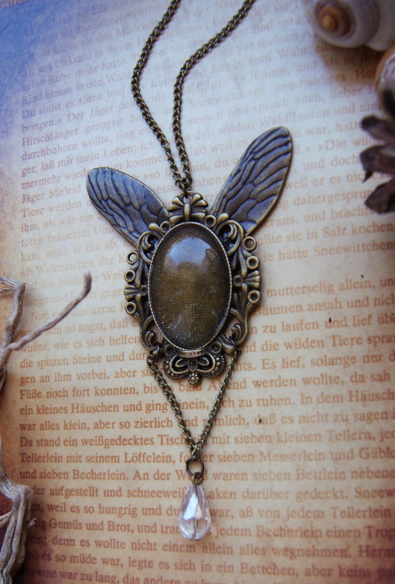 Puede incluir: Un collar de bronce con un gran colgante ovalado que presenta un cabuj&oacute;n de vidrio transparente y dos alas detalladas. El collar est&aacute; sobre un libro con texto en alem&aacute;n.