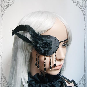 May include: A black leather eye patch with a black rose and feather embellishment. The eye patch has a beaded chain with black teardrop-shaped beads.