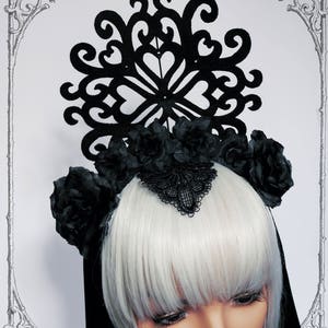 Puede incluir: Un tocado de estilo g&oacute;tico negro con un gran dise&ntilde;o de corte intrincado y rosas negras. El tocado lo lleva una persona con el pelo blanco.