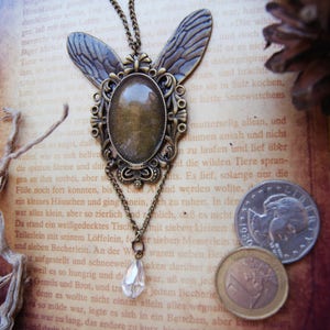 Puede incluir: Un collar de bronce con un colgante de mariposa con un cabuj&oacute;n de cristal transparente. El collar est&aacute; sobre un libro con texto en alem&aacute;n. El colgante est&aacute; adornado con intrincados detalles.