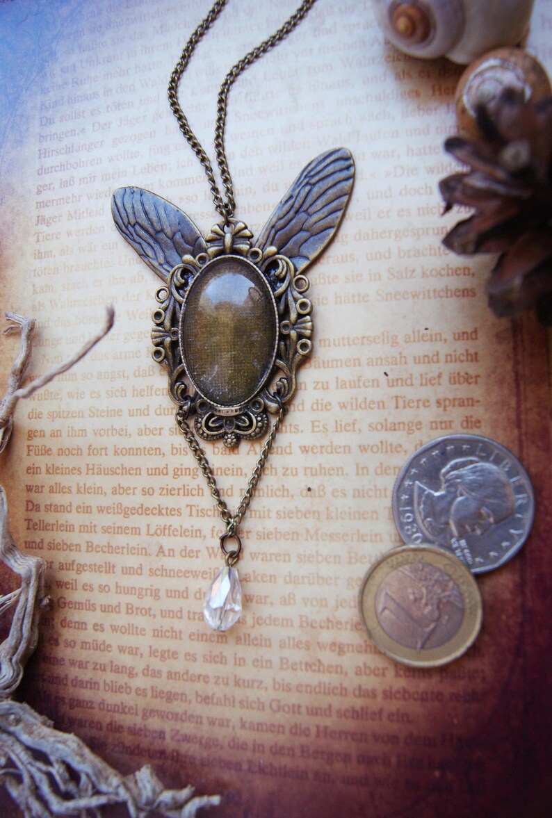 Puede incluir: Un collar de bronce con un colgante de mariposa con un cabuj&oacute;n de cristal transparente. El collar est&aacute; sobre un libro con texto en alem&aacute;n. El colgante est&aacute; adornado con intrincados detalles.