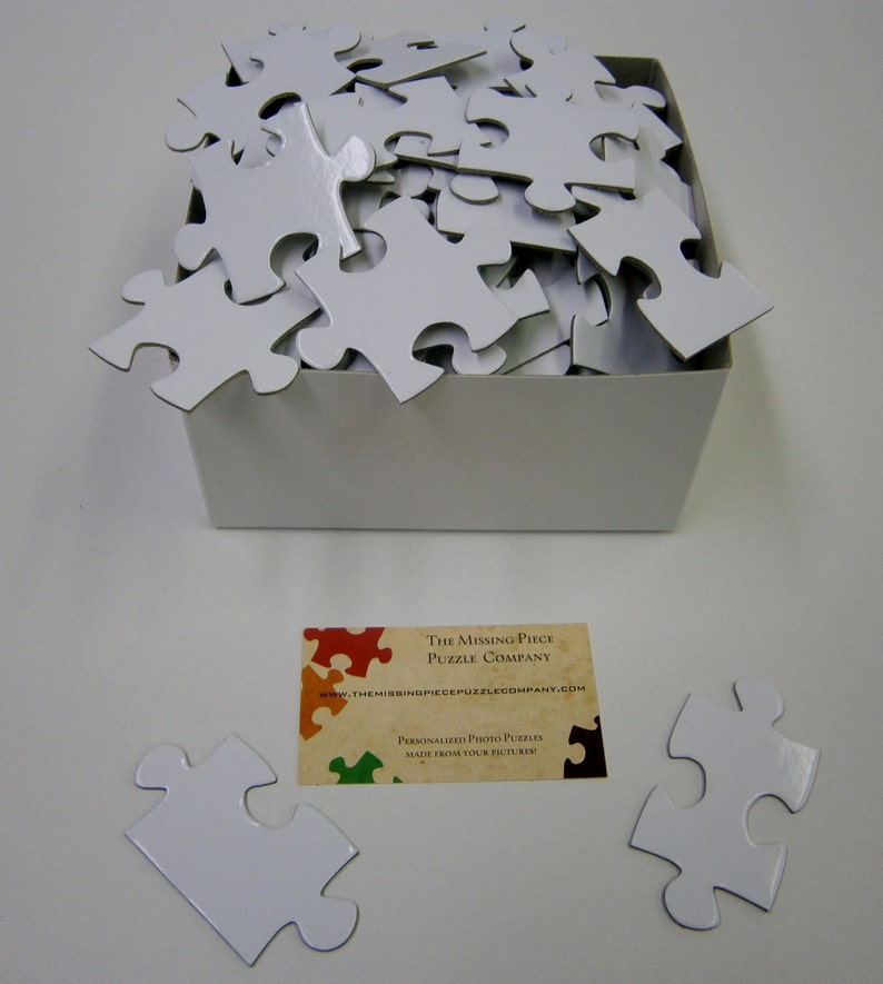 Puede incluir: Una caja blanca llena de piezas de rompecabezas blancas. La caja est&aacute; sobre una superficie blanca. Hay una tarjeta de visita en la superficie que dice "The Missing Piece Puzzle Company" y "Personalized Photo Puzzles Made From Your Pictures".