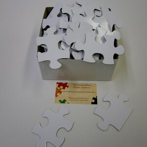 Puede incluir: Una caja blanca llena de piezas de rompecabezas blancas en blanco. La caja est&aacute; sentada sobre una superficie blanca. Una tarjeta de presentaci&oacute;n para "The Missing Piece Puzzle Company" est&aacute; en primer plano. La tarjeta tiene un dise&ntilde;o de rompecabezas colorido y el texto "Personalized Photo Puzzles Made from Your Pictures".