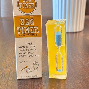 May include: A vintage egg timer with a yellow and white box, and a clear yellow hourglass. The box reads "EGG TIMER 3 MINUTE TIMER" and lists uses like "MORNING EGGS" and "PHONE CALLS." The hourglass has blue sand.