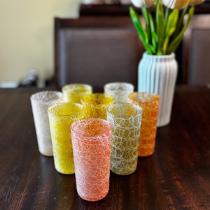 May include: A collection of eight cylindrical drinking glasses featuring a textured, white spaghetti-style web pattern over translucent colors including orange, yellow, green, and off-white. The glasses are arranged in a cluster on a dark surface.