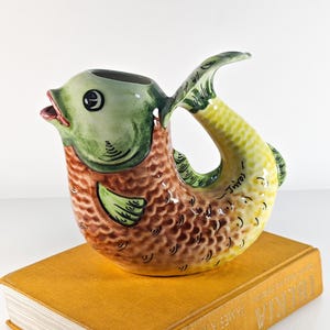 May include: A ceramic fish-shaped pitcher with a green head, brown body, and yellow tail, resting on a mustard-colored book. The pitcher has an open mouth and a handle formed by the tail. The book's title is partially visible.