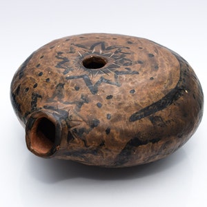 Può includere: Un vaso di terracotta marrone con disegni dipinti in nero. Il vaso ha un corpo rotondo e una piccola apertura nella parte superiore. I disegni includono un motivo a stella e punti.