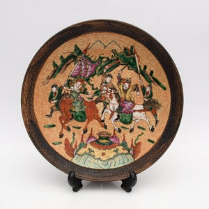 Puede incluir: Un plato de cerámica decorativo con una ilustración detallada de una escena de batalla con figuras a caballo, montañas y banderas. El plato tiene un borde marrón y un fondo beige con un efecto craquelado. El plato se exhibe sobre un soporte negro.