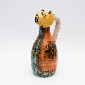 May include: A ceramic pitcher shaped like a cat with a yellow head, orange body, and green and blue floral designs. The pitcher has a white handle.