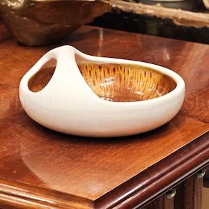 Peut inclure: Un bol en céramique blanc avec une poignée unique. L'intérieur présente un motif texturé marron et doré. Le bol est posé sur une surface en bois poli, mettant en valeur sa forme élégante. Un objet décoratif.