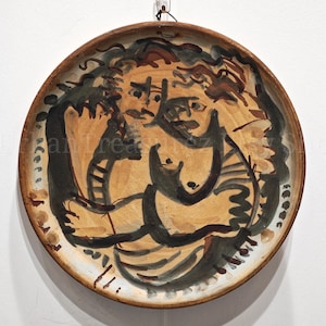 May include: A round, hand-painted ceramic plate with a brown rim and a central abstract design. The artwork features figures in shades of brown, black, and beige. The plate is hanging against a white wall.