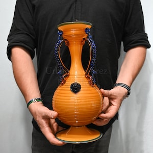 Puede incluir: Un jarrón de vidrio grande, de color naranja, con detalles azules y marrones. El jarrón tiene dos asas y una base redonda. Presenta un diseño decorativo de cabeza de león.