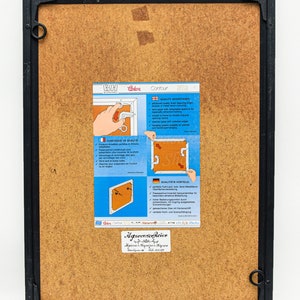 May include: A black picture frame with a brown cardboard backing. The frame has a metal ring on the top right corner. A white label with the text "Agrocorico Reior" is attached to the bottom right corner of the frame. A white label with the text "Contour" is attached to the frame. The label has a diagram of a picture frame with a metal ring attached to the frame. The label has text in German, French, and English describing the features of the frame.