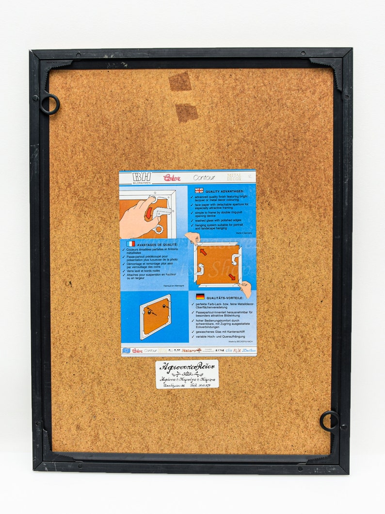 May include: A black picture frame with a brown cardboard backing. The frame has a metal ring on the top right corner. A white label with the text "Agrocorico Reior" is attached to the bottom right corner of the frame. A white label with the text "Contour" is attached to the frame. The label has a diagram of a picture frame with a metal ring attached to the frame. The label has text in German, French, and English describing the features of the frame.