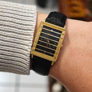 Puede incluir: Reloj Orient rectangular dorado con esfera negra y un diamante. El reloj tiene barras horizontales doradas y la palabra "ORIENT" está impresa en la esfera. Está sujeto a una correa de cuero negro.