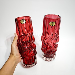 May include: Two ruby red Bohemian glass vases with a textured, wavy design. Each vase has a clear, textured base and a gold and black label that reads "Bohemia". The vases are tall and cylindrical, with a unique, organic shape.