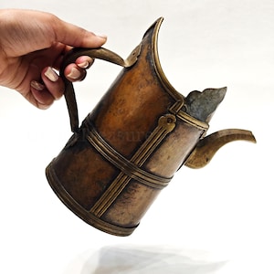 May include: A cylindrical brass pitcher with a weathered, dark patina finish. The vessel features a curved handle, a wide spout, and decorative vertical and horizontal brass bands encircling the body, creating a structured, metallic appearance.