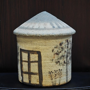 Op de afbeelding: Een cilindrische keramische pot met een conisch deksel, voorzien van een beige getextureerd oppervlak versierd met handgeschilderde donkerbruine illustraties van een rechthoekig raam, een boom en kleine bloemen, bekroond met een grijs gespikkeld dak.