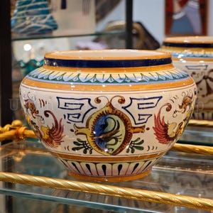 Peut inclure: Un bol en céramique rond à base blanche, orné de motifs décoratifs peints à la main en orange, bleu, vert et rouge. Le design comprend des figures stylisées rappelant des oiseaux, des motifs géométriques et un emblème ovale central bleu.