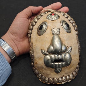 May include: An ornate, cream-colored mask with silver metal accents. The mask features a prominent nose, eyes, and mouth, with decorative elements around the edges. A small red stone is set at the top. The mask is held by a hand.