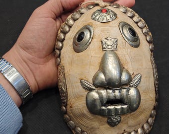 May include: An ornate, cream-colored mask with silver metal accents. The mask features a prominent nose, eyes, and mouth, with decorative elements around the edges. A small red stone is set at the top. The mask is held by a hand.