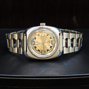 Könnte beinhalten: Eine silberfarbene Armbanduhr mit metallisch goldenem Zifferblatt, einem Datumsfenster auf der Drei-Uhr-Position und dem Aufdruck "WATERPROOF 25 METRES". Die Uhr verfügt über ein Edelstahl-Gliederarmband mit ausgeschnittenen Details.