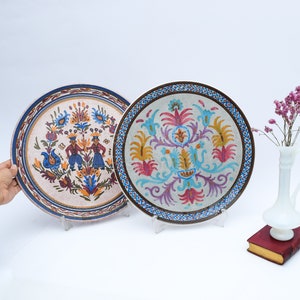 May include: Two decorative ceramic plates with colorful floral and folk art designs. The plates are on a white surface with a white vase and dried flowers.