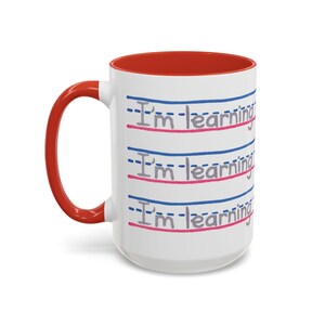 May include: White ceramic mug with a red interior and handle. The mug features the text "I'm learning" in a handwritten style, repeated three times with blue and pink lines, creating a school theme.