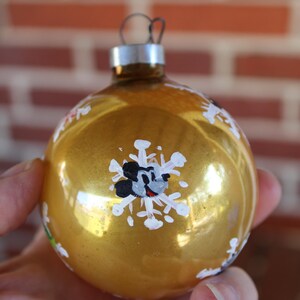 May include: Gold Christmas ornament with a painted Mickey Mouse design on a white snowflake. The ornament is decorated with white snowflakes and other painted details.